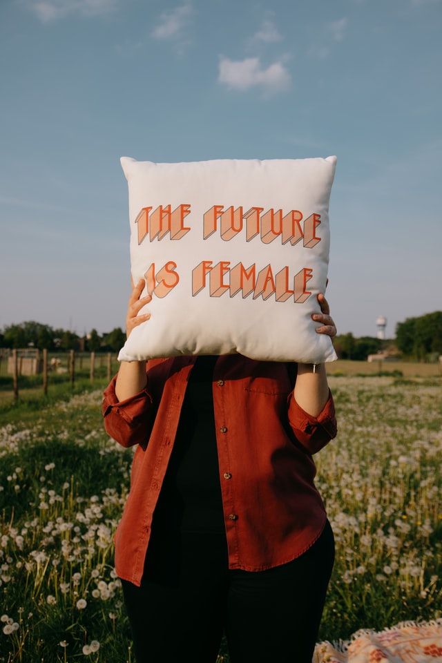 Woman in red shirt standingn with a pillow covering her face. On the pillow are the words the future is female